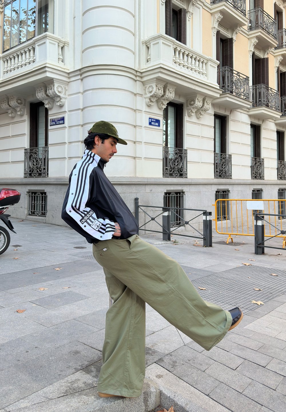 Black zip-up jacket with white stripes, olive green wide-legged trousers, and a green cap. Standing on a street with buildings in the background.