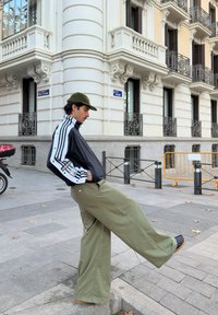 Black zip-up jacket with white stripes, olive green wide-legged trousers, and a green cap. Standing on a street with buildings in the background.