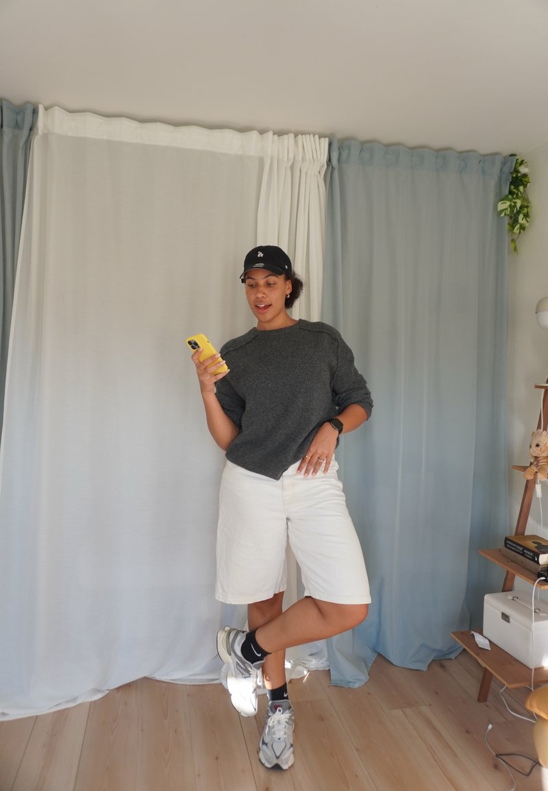 Grigia maglietta con dettagli a coste, pantaloni corti bianchi e sneakers. In piedi su un pavimento in legno, tiene un telefono giallo, davanti a tende chiare.
