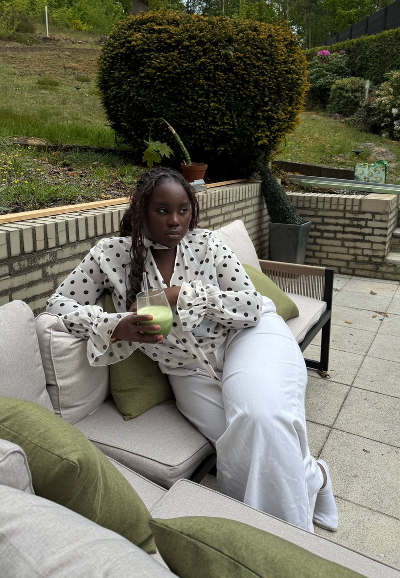 Femme en blouse blanche à pois blancs et pantalon blanc assise sur un canapé extérieur tenant une boisson verte, avec de la verdure et un mur en briques en arrière-plan.