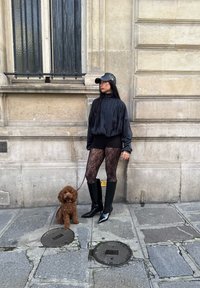 Kvinna i svart outfit, spetsstrumpbyxor och stövlar håller i koppel till liten brun krullig hund på stadens trottoar vid stenbyggnad.