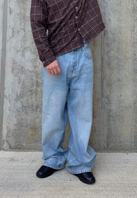 Hellblaue, weit geschnittene Jeans mit einem ausgewaschenen Finish, getragen zu einem dunkelbraunen Hemd mit Knöpfen und Karomuster. Schwarze Schuhe vervollständigen das Outfit.