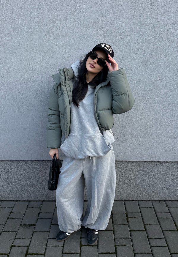 Chaqueta tipo puffer en verde oliva, sudadera gris y pantalones de chándal. Gorra negra con logo y gafas de sol. Bolso negro y zapatos oscuros.