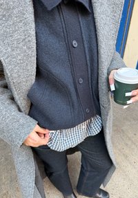 Personne tenant une tasse à café à emporter verte, portant un manteau gris, un pull noir boutonné, une chemise à carreaux, un pantalon noir et des ongles longs avec des extrémités blanches.
