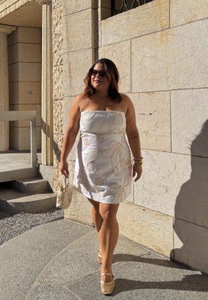 Mujer vestida con un vestido blanco sin mangas y con patrones florales beiges, caminando junto a una pared de piedra, sosteniendo un bolso blanco texturizado y luciendo gafas de sol.