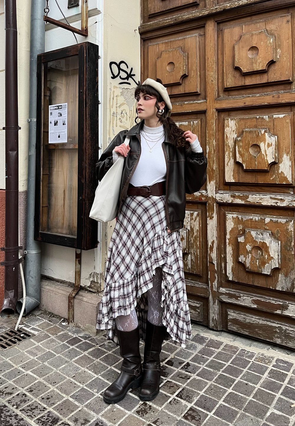 Veste en cuir marron sur une chemise blanche, associée à une jupe à carreaux avec volants et des bottes noires montant jusqu'aux genoux. Béret beige et un sac cabas blanc.