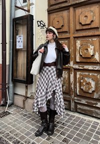 Veste en cuir marron sur une chemise blanche, associée à une jupe à carreaux avec volants et des bottes noires montant jusqu'aux genoux. Béret beige et un sac cabas blanc.
