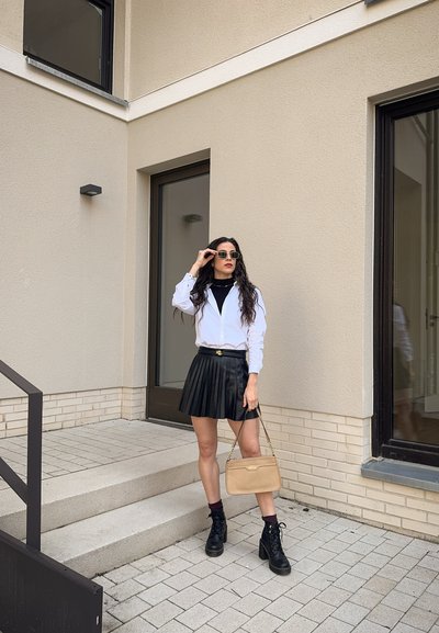 Camisa blanca de botones, falda negra con plisados, botas de tobillo negras con cordones y un bolso beige. El entorno presenta una arquitectura moderna con escalones de piedra.