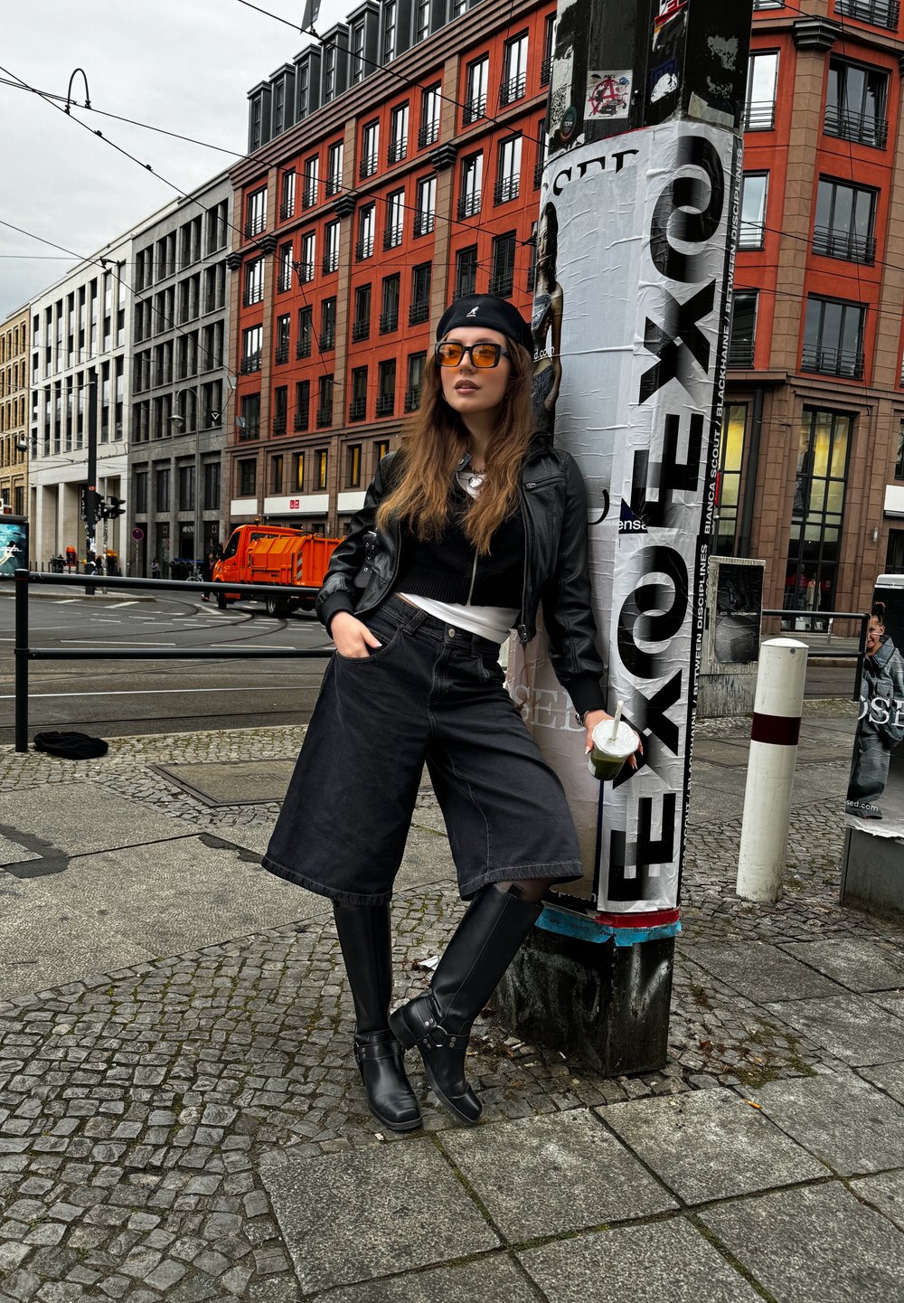 Μαύρο δερμάτινο μπουφάν πάνω από ένα μαύρο cropped πουλόβερ, φαρδιά μαύρα τζιν σορτς και μπότες μέχρι το γόνατο σε μαύρο χρώμα. Τα αξεσουάρ περιλαμβάνουν γυαλιά ηλίου και ένα ποτό.