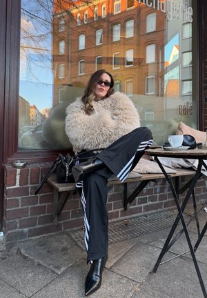 Mujer con gafas de sol y chaqueta esponjosa sentada con las piernas cruzadas en un banco de café al aire libre, con una taza de café y un bolso en la mesa cerca.