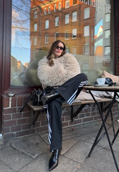 Mujer con gafas de sol y chaqueta esponjosa sentada con las piernas cruzadas en un banco de café al aire libre, con una taza de café y un bolso en la mesa cerca.