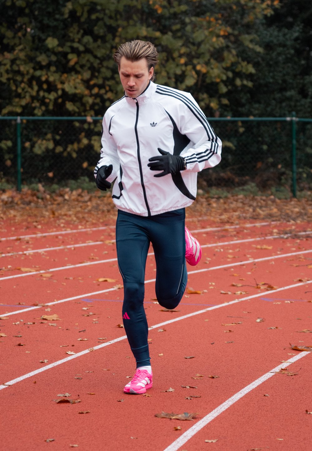 Coureur masculin en veste blanche, leggings bleu marine et chaussures roses, en train de jogger sur une piste rouge extérieure couverte de feuilles tombées.