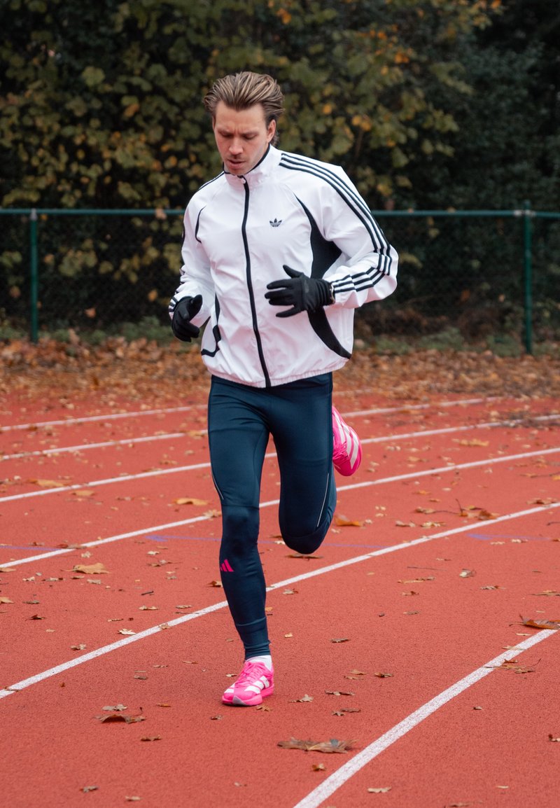Un coureur masculin en veste blanche, leggings bleu marine et chaussures roses fait du jogging sur une piste extérieure rouge couverte de feuilles tombées.