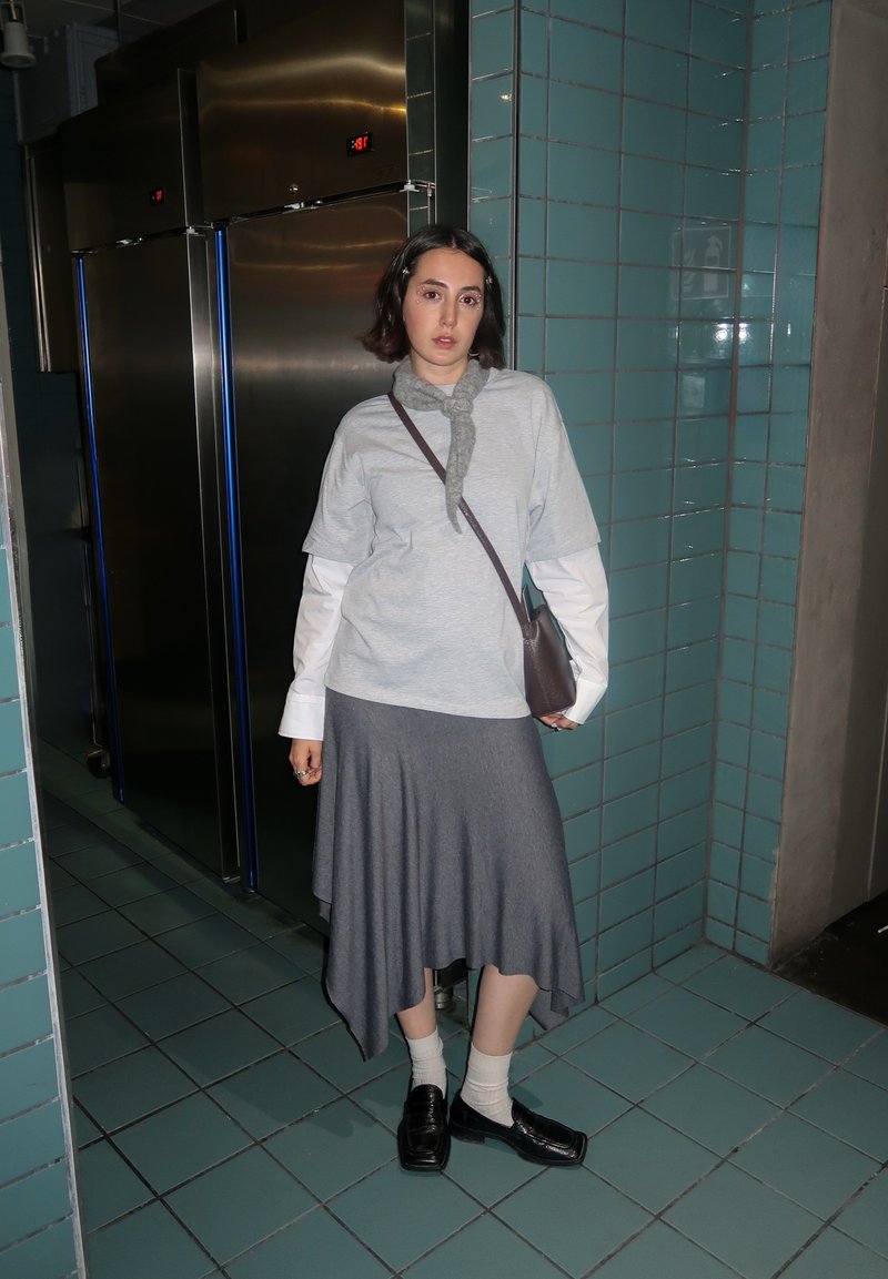Grey T-shirt layered over a white long-sleeve shirt, paired with a grey asymmetrical skirt. Black loafers and light socks complete the look.