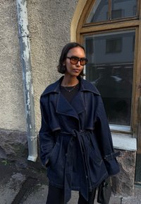 Denim jakna v temno modri barvi s sproščenim krojem, velikim ovratnikom, pasom na vezavo in stranskimi žepi. Nosi se preko črne majice z teksturiranim zaključkom.