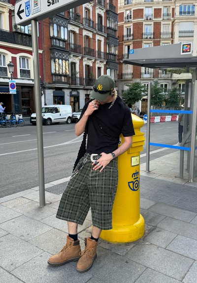 Camisa negra de manga corta, pantalones cortos de cuadros verdes hasta la rodilla, botas marrones con cordones. Los accesorios incluyen un cinturón negro y una gorra verde con un logotipo.