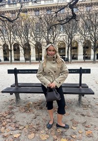 Beige trenssitakki leveillä kauluksilla ja napeilla, mustat lyhyet housut, mustat ballerinat ja tummanruskea käsilaukku. Taustalla puita ja puiston penkki.