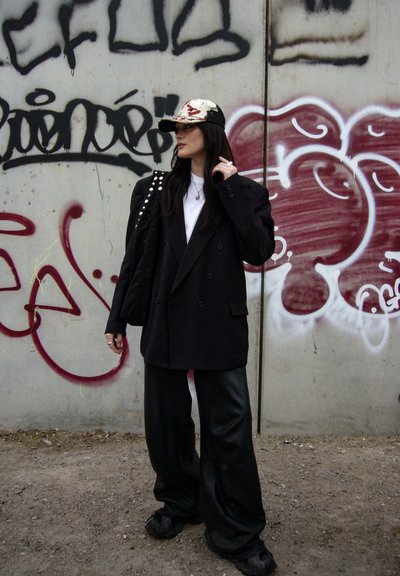 Mujer con una blazer negra extragrande, pantalones anchos, gorra estampada y lleva un bolso negro con tachuelas, de pie frente a una pared de hormigón cubierta de grafitis.