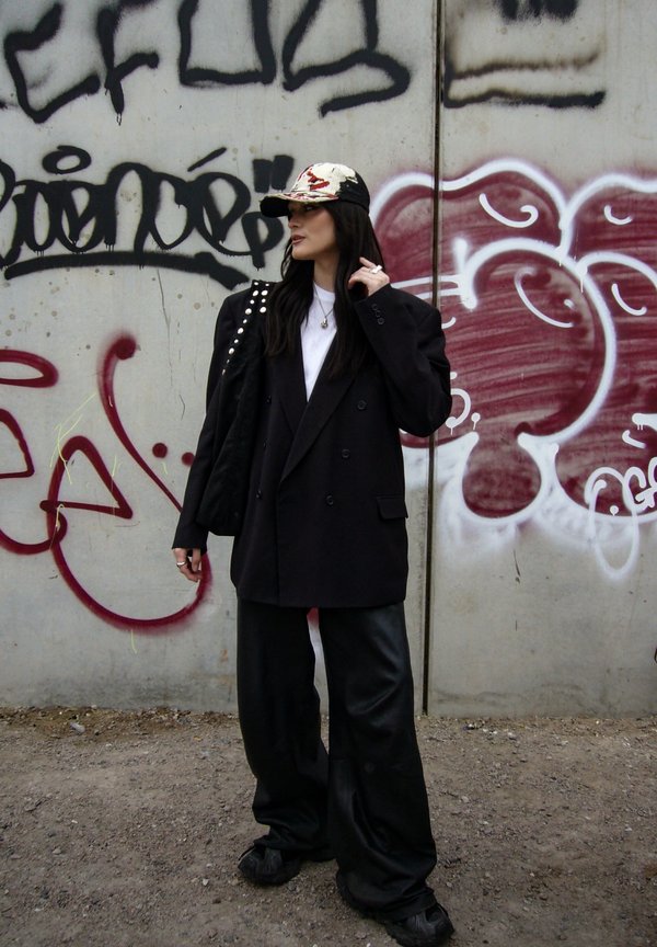 Žena v čiernom oversized saku, širokých nohaviciach, vzorovanej šiltovke a nesúca čiernu kabelku s cvokmi, stojaca pri betónovej stene pokrytej graffiti.