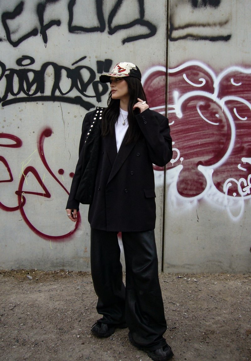 Femme portant un blazer noir oversized, un pantalon large, une casquette à motifs et portant un sac noir clouté, debout devant un mur en béton couvert de graffitis.