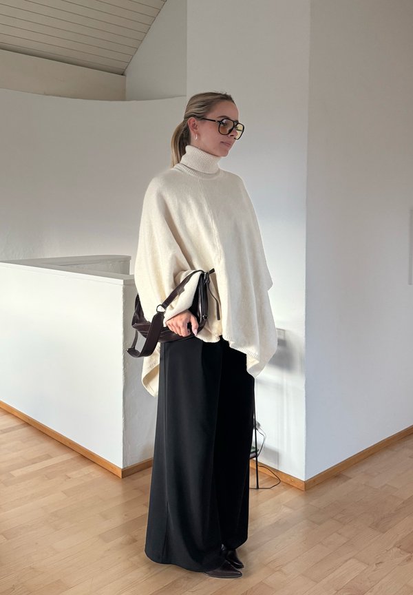 Mujer con un poncho de cuello alto blanco, pantalones anchos negros, gafas y sosteniendo un bolso negro en el interior sobre un suelo de madera.