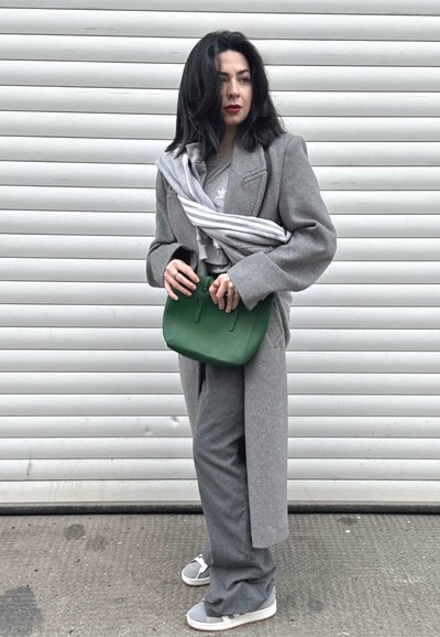 Abrigo largo gris sobre un conjunto gris, acompañado de un bolso de cuero verde. Presenta una parte superior gris en capas con rayas y zapatillas gris.