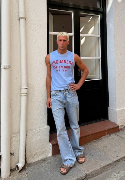 Camiseta sin mangas de color azul claro con texto rojo, jeans azul deslavado y sandalias marrones; de pie en una puerta con marco negro y pared blanca.