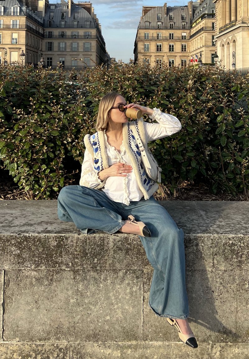 Ένα άτομο κάθεται σε μια πέτρινη προεξοχή φορώντας μια λευκή μπλούζα, ένα γαλάζιο κεντημένο γιλέκο και φαρδιές τζιν παντελόνες. Είναι ορατά μαύρα και γυμνά τακούνια.