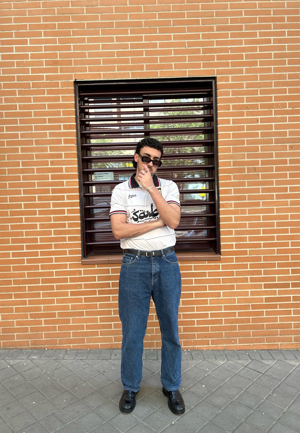 Polo alb cu mânecă scurtă, cu accente negre și roșii, blugi din denim albastru și pantofi negri, stând lângă un perete de cărămidă și o fereastră.