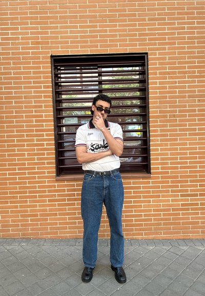 Polo blanco de manga corta con acentos en negro y rojo, jeans de mezclilla azules y zapatos negros, de pie contra una pared de ladrillo y una ventana.