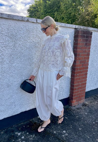 Mujer con el pelo rubio recogido en una coleta lleva una blusa blanca de encaje, pantalones blancos sueltos, chanclas negras, gafas de sol y sostiene un pequeño bolso negro mientras camina junto a una pared.
