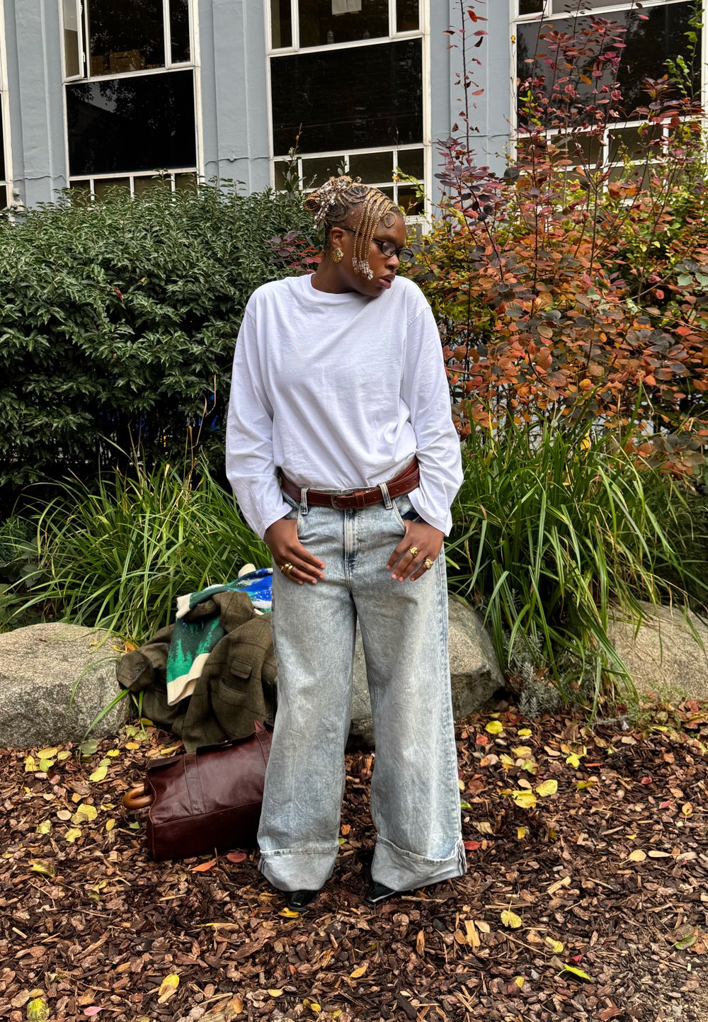 Hosszú ujjú fehér ing, laza, világoskék farmer, barna öv és fekete cipő. A háttérben zöld bokrok és barna mulcs látható.