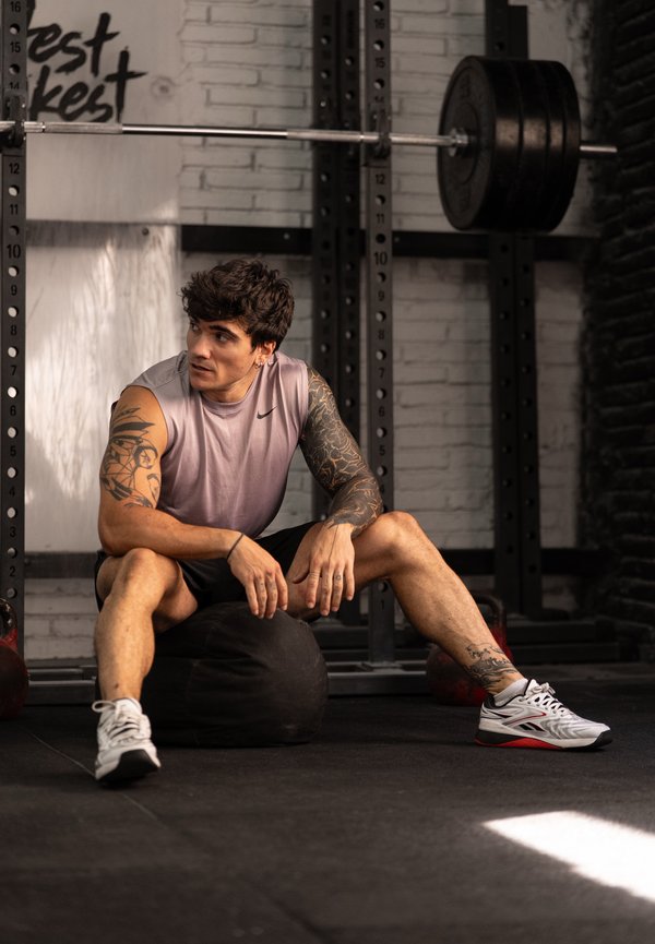 Mand med tatoveringer, der hviler på en sort medicinbold inde i et fitnesscenter med en vægtstang og vægtstativ bag ham.