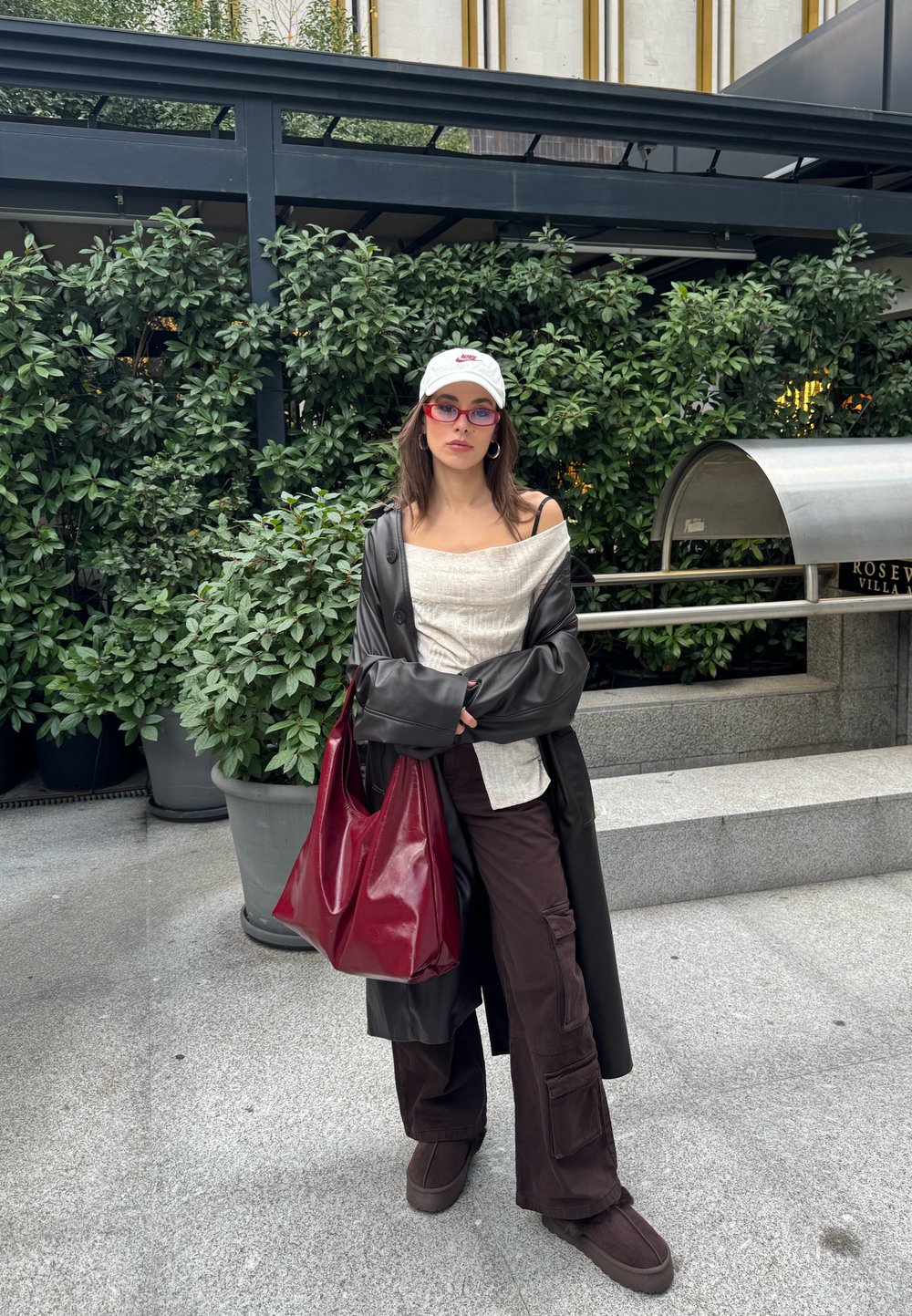 Femme portant un manteau en cuir noir, un haut blanc décolleté, un pantalon cargo marron, une casquette blanche, des lunettes rouges, tenant un grand sac rouge, debout près des plantes.