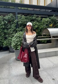 Femme portant un manteau en cuir noir, un haut blanc décolleté, un pantalon cargo marron, une casquette blanche, des lunettes rouges, tenant un grand sac rouge, debout près des plantes.