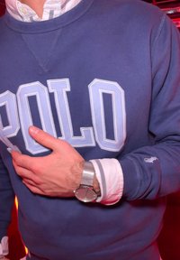 Mornarsko moder pulover z belim napisom "POLO", prekrit nad beli in modri prugasti srajco s kragenom. Na zapestju je nosjena srebrna mrežasta ura.