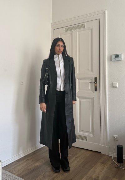 Mujer de pie en el interior vistiendo un abrigo largo de cuero negro, camiseta blanca con cremallera, pantalones negros y zapatos negros, junto a una puerta blanca cerrada.
