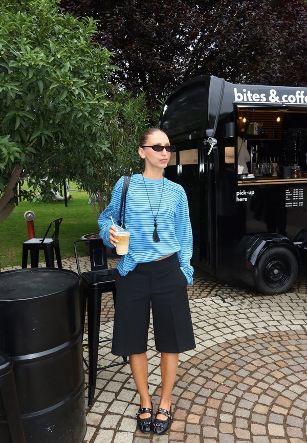 Frau mit Sonnenbrille, die Eiskaffee hält, steht neben einem schwarzen Kaffeewagen mit Außenbestuhlung und Grünpflanzen im Hintergrund.