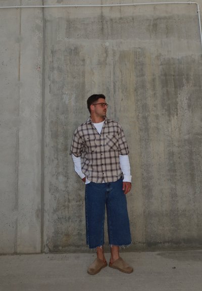 Hombre con gafas, camisa de cuadros sobre camiseta blanca de manga larga, vaqueros anchos recortados y zapatos beige sin cordones, apoyado contra una pared de hormigón.