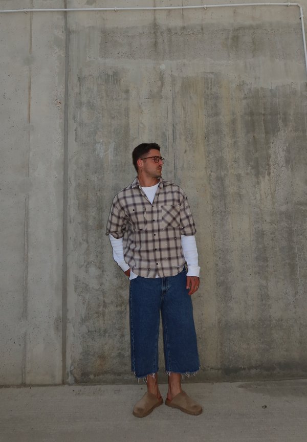 Mann med briller, rutete skjorte over hvit langermet genser, vide korte jeans og beige slip-on sko står mot en betongvegg.