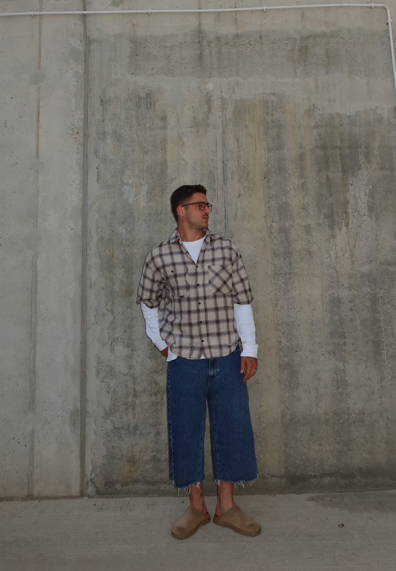Mann mit Brille, kariertem Hemd über weißen Langarmshirt, weit geschnittene kurze Jeans und beigen Slippern steht an einer Betonwand.