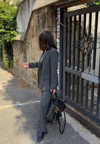 Szürke kockás öltöny strukturált blézerrel és bőszárú nadrággal. Fekete kézitáska és bőrcipő. Kőfal és fémkapu a háttérben.