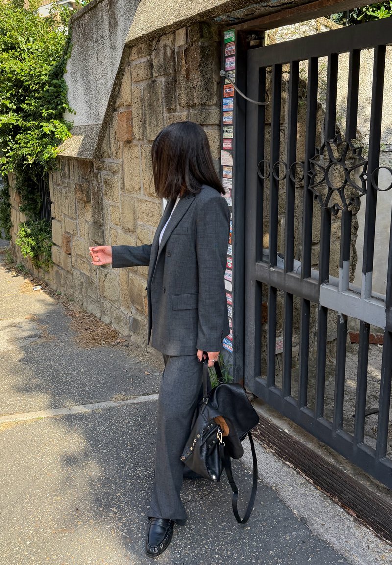 Fato xadrez cinzento com blazer estruturado e calças de perna larga. Mala preta e sapatos de couro. Parede de pedra e portão metálico ao fundo.
