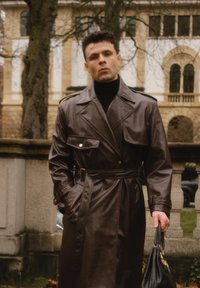 Abrigo trench de cuero marrón con cintura ceñida con cinturón, grandes bolsillos con solapa y cuello ancho. Lleva un suéter de cuello alto negro y sostiene un bolso negro.