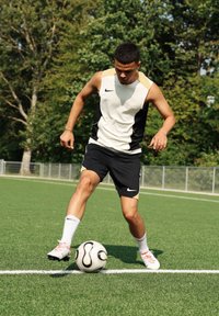 Utøver i en bærekraftig beige og svart Nike-top uten ermer med svarte shorts, dribler en svart og hvit fotball på kunstgress.