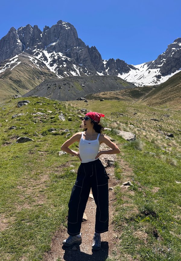 Jeune femme en chaussures de randonnée, pantalon noir, débardeur blanc et bandana rouge, debout sur un sentier de montagne avec des sommets rocheux et des plaques de neige en arrière-plan.