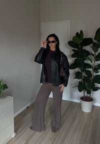 Žena v černé kožené bundě, tmavých slunečních brýlích a hnědých širokých kalhotách stojí uvnitř vedle květináče a bílé komody.