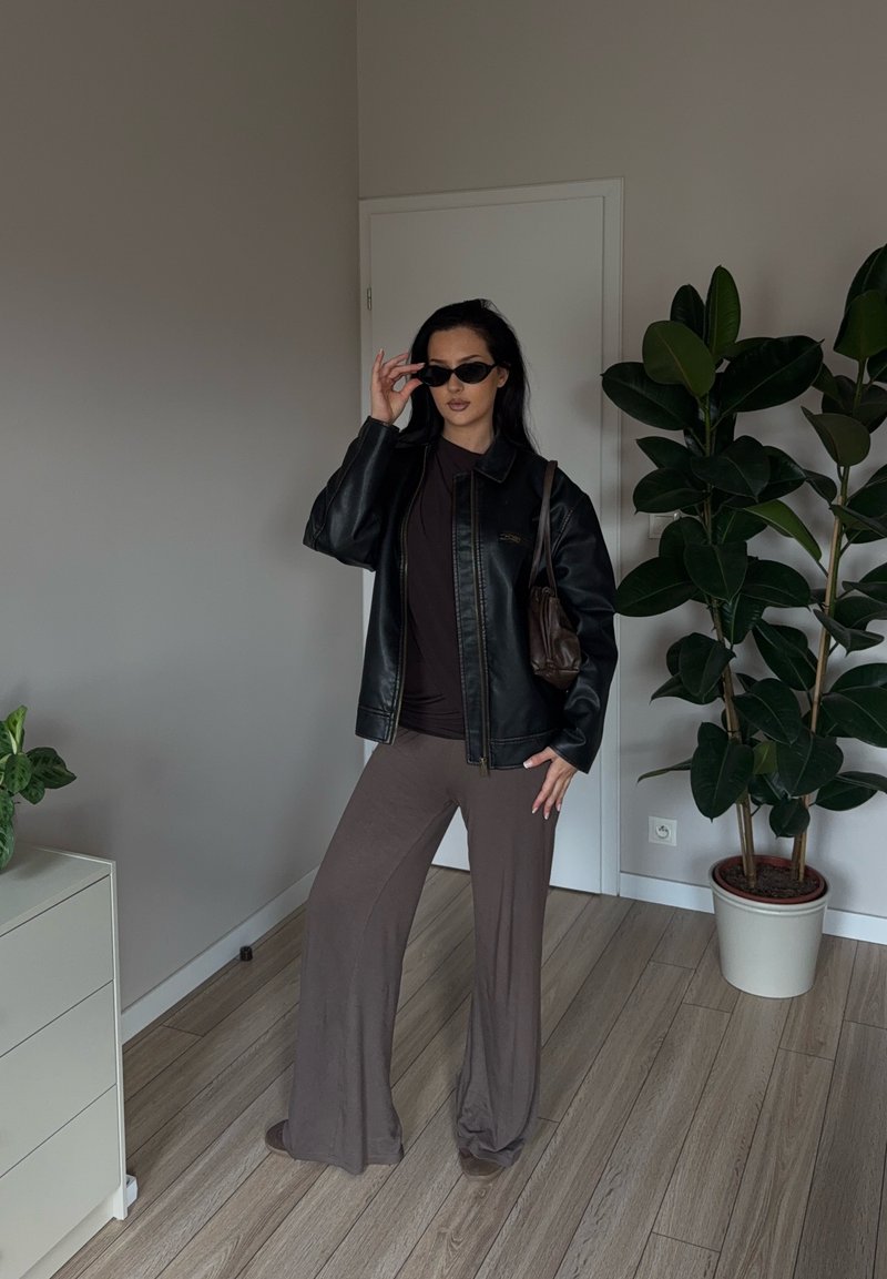 Donna che indossa una giacca di pelle nera, occhiali da sole scuri, pantaloni larghi marroni, in piedi all'interno accanto a una pianta in vaso e a un comò bianco.