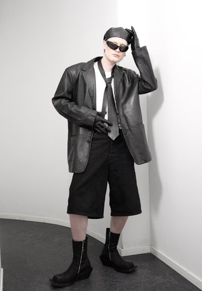 Persona con chaqueta de cuero negra, guantes, gorra, gafas de sol, corbata, pantalones cortos anchos hasta la rodilla y botas negras, de pie en una esquina contra paredes blancas.