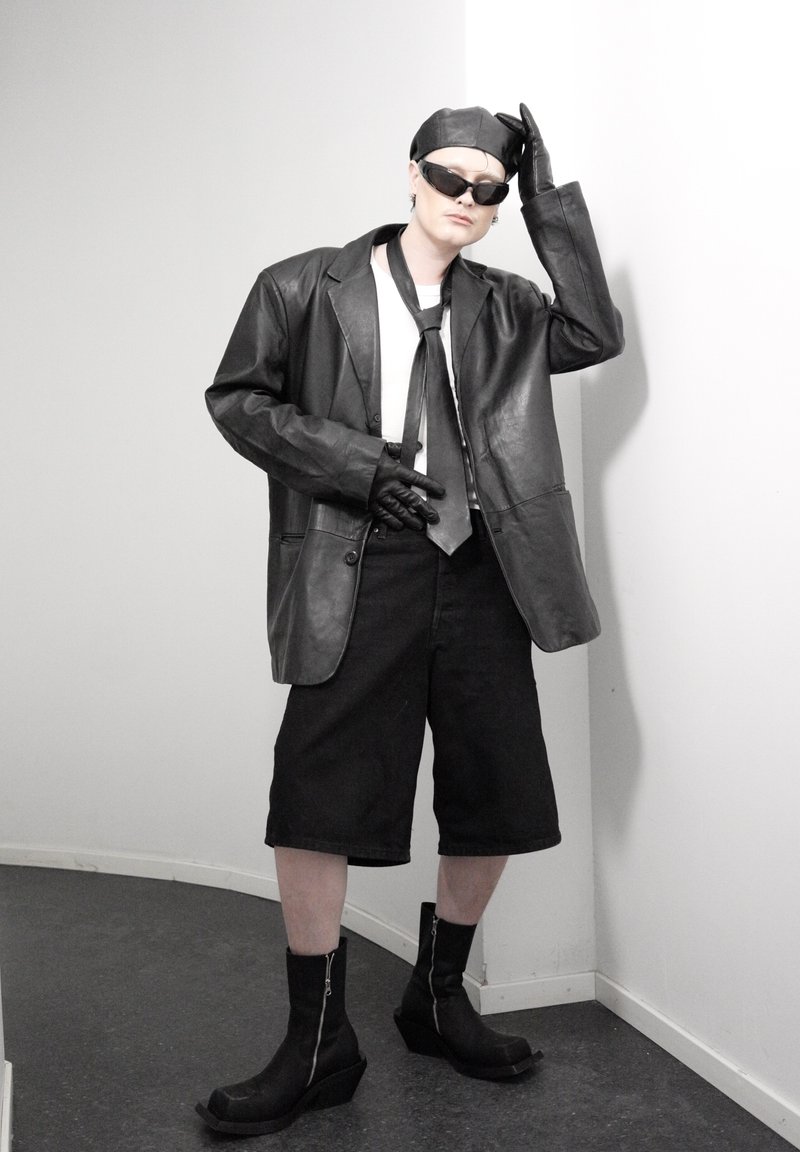 Personne portant une veste en cuir noire, des gants, une casquette, des lunettes de soleil, une cravate, un short ample arrivant aux genoux et des bottes noires, debout dans un coin contre des murs blancs.